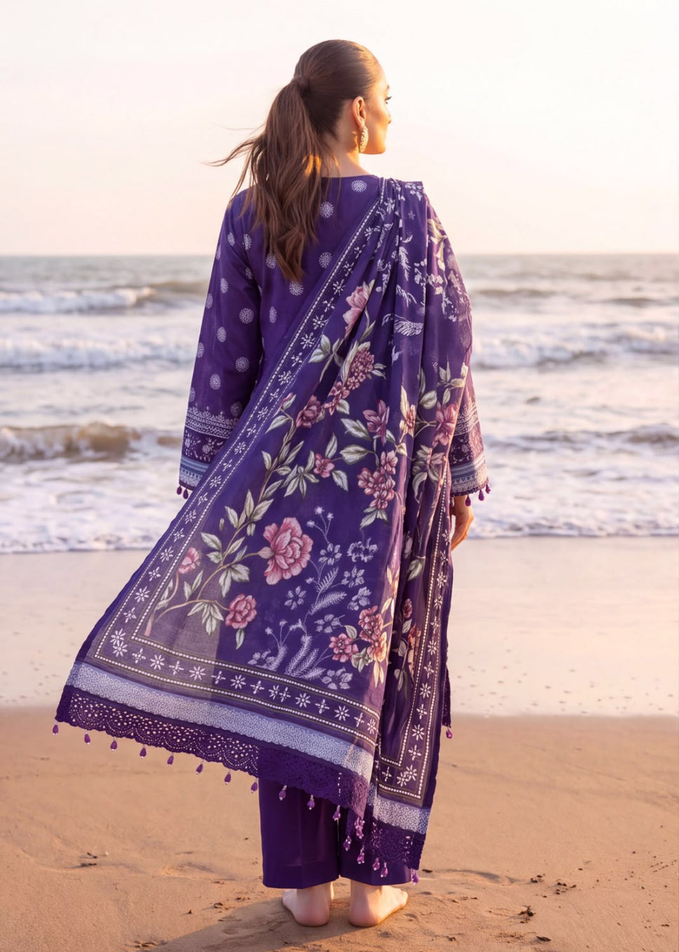 A woman stands on a sandy beach facing the ocean, wearing a purple traditional outfit with floral prints and a matching dupatta.