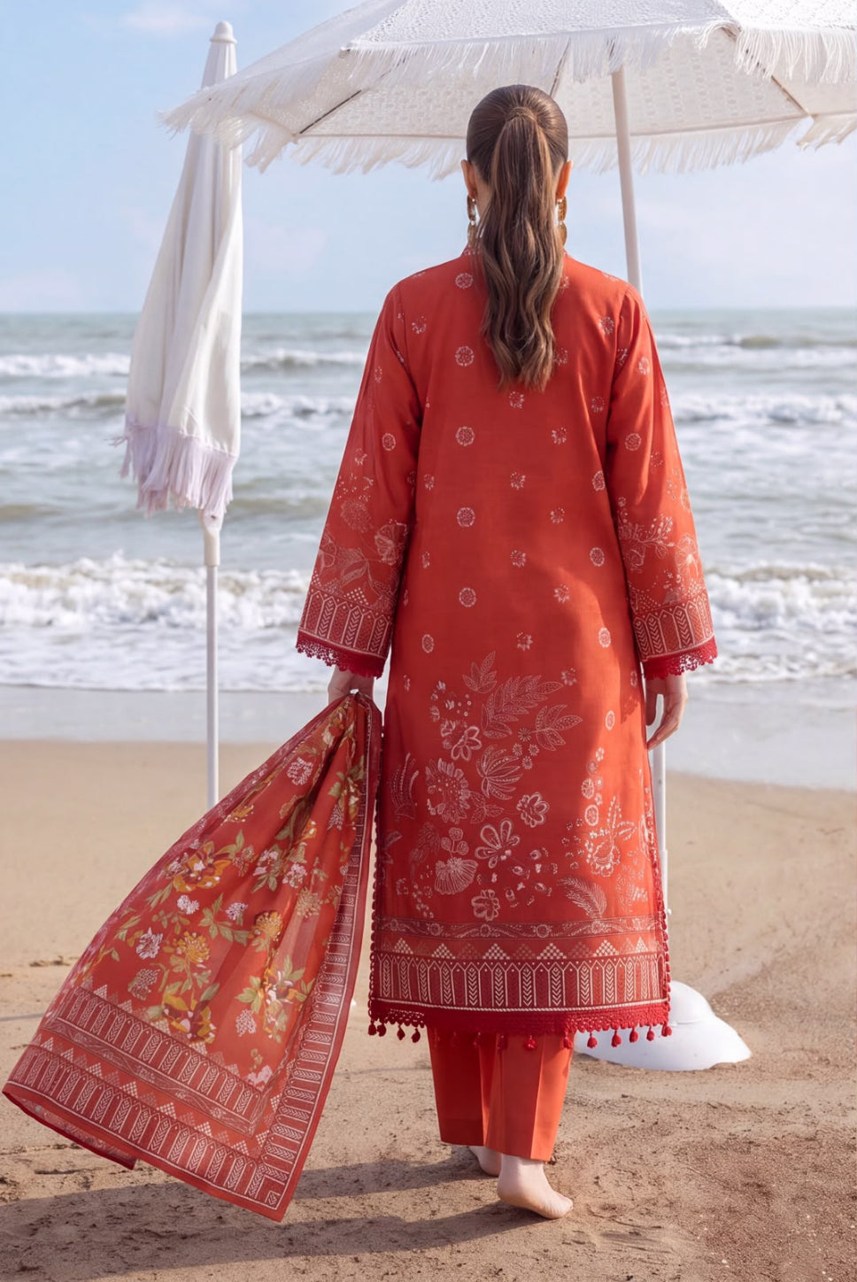 A woman stands on a sandy beach facing the ocean, wearing a rust-orange embroidered kurta with matching pants and holding a coordinating dupatta. A white parasol is open behind her.
