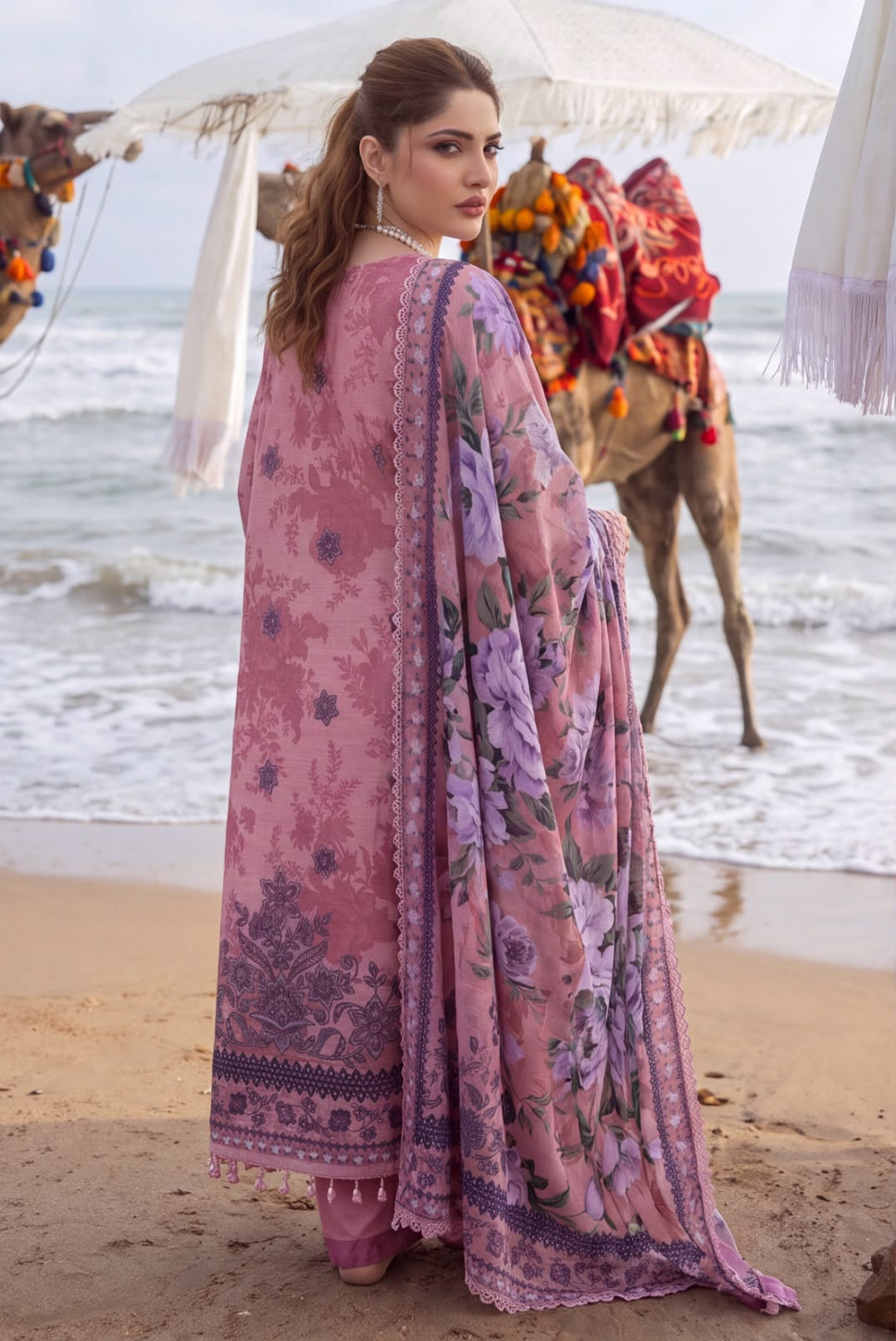 A woman stands on a sandy beach facing away from the camera, wearing a pink floral traditional outfit with a matching dupatta. Camels with colorful saddles and white umbrellas are visible in the background near the ocean.