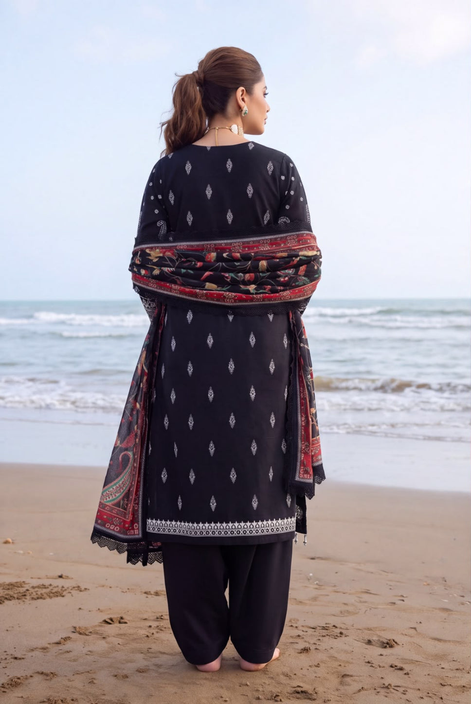 A woman stands on a sandy beach facing the ocean, wearing a black three-piece salwar kameez suit with small white geometric patterns, black trousers, and a red and black patterned dupatta.