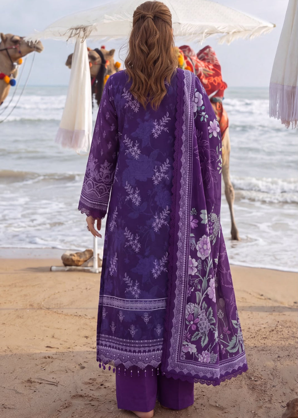 A woman stands on a sandy beach facing the ocean, wearing a long purple embroidered traditional outfit with matching pants and a floral dupatta. Camels and white umbrellas are visible in the background near the shoreline.