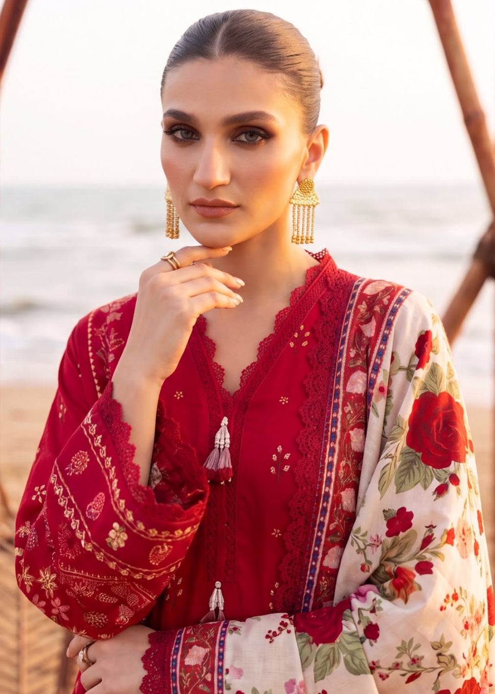 A woman posing outdoors near the beach, wearing a traditional red and cream floral embroidered kurta with gold-toned earrings.