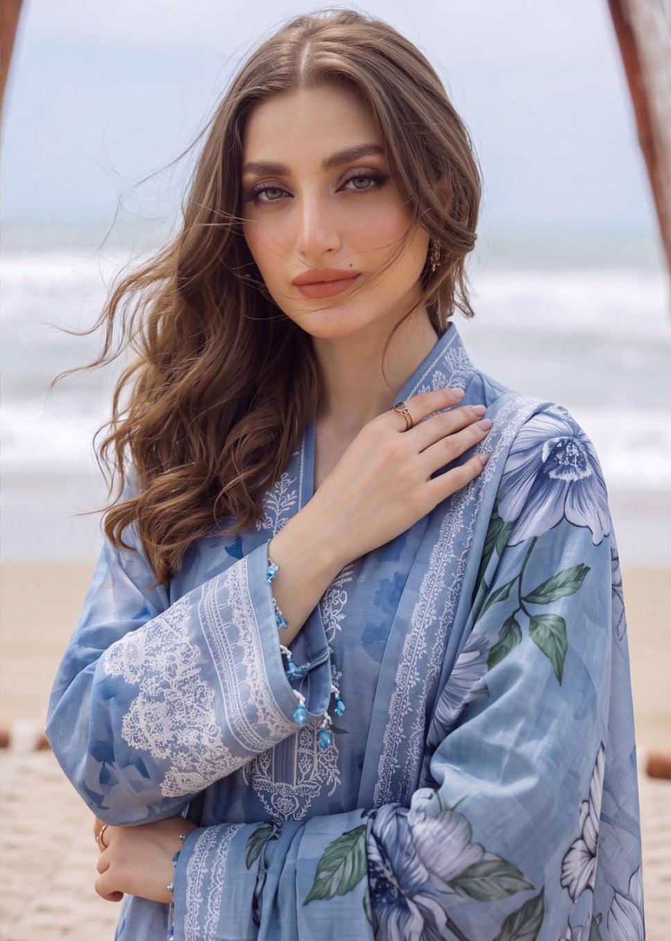 A woman wearing a light blue floral printed kaftan-style dress with white embroidery, standing on a sandy beach with the ocean in the background.