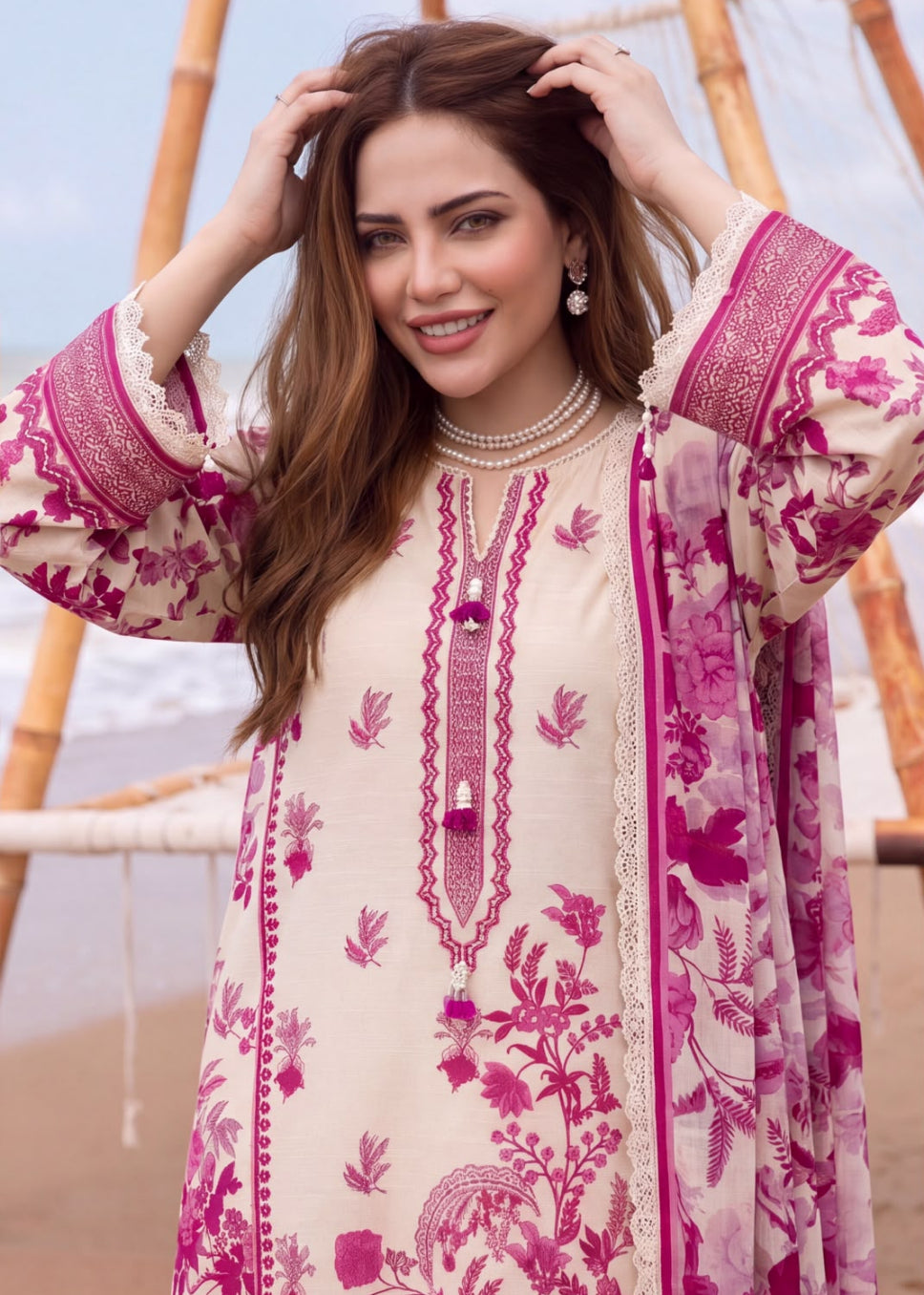 A woman wearing a beige and magenta floral printed traditional outfit with a matching sheer dupatta, standing in front of a bamboo structure.