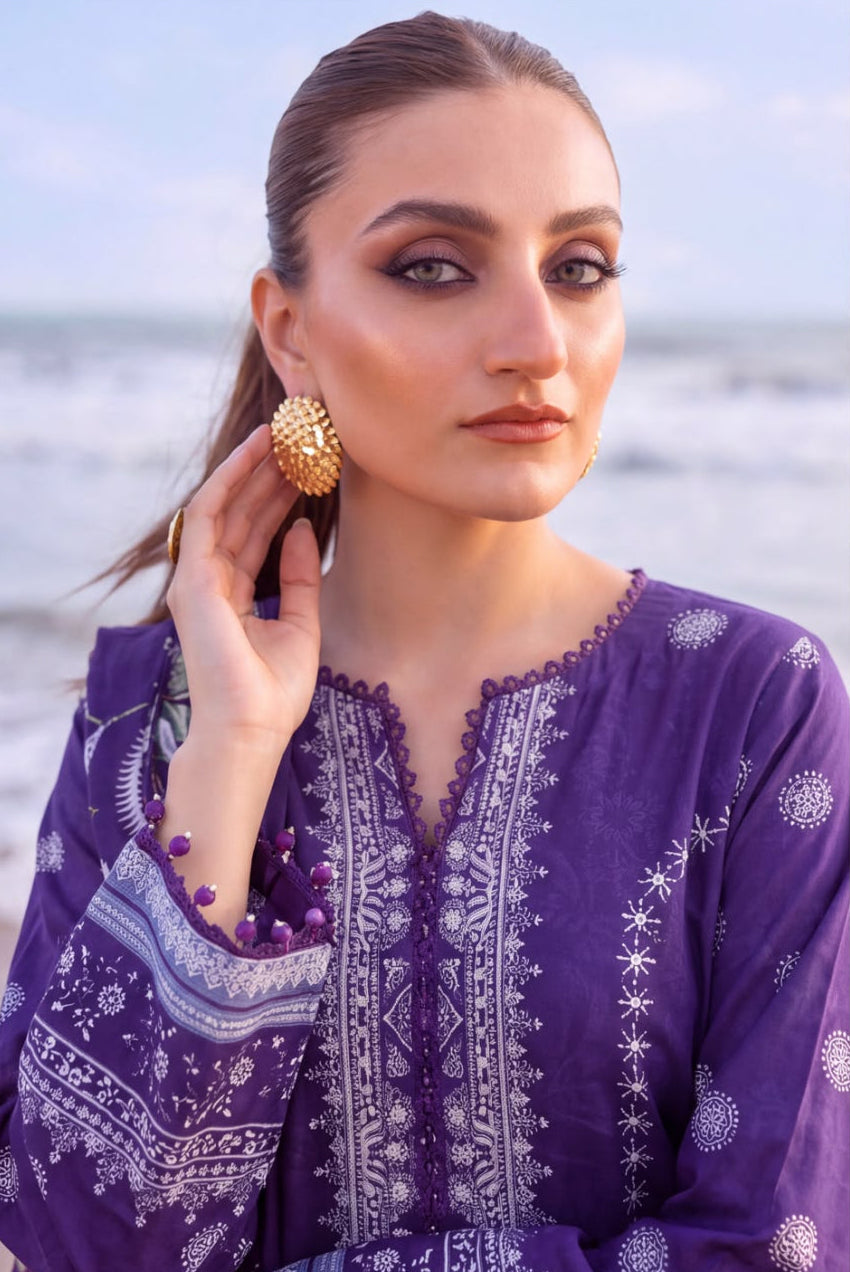 A woman wearing a purple embroidered tunic top with white patterns, standing outdoors near the ocean. She is also wearing a chunky gold statement earring and a beaded bracelet.
