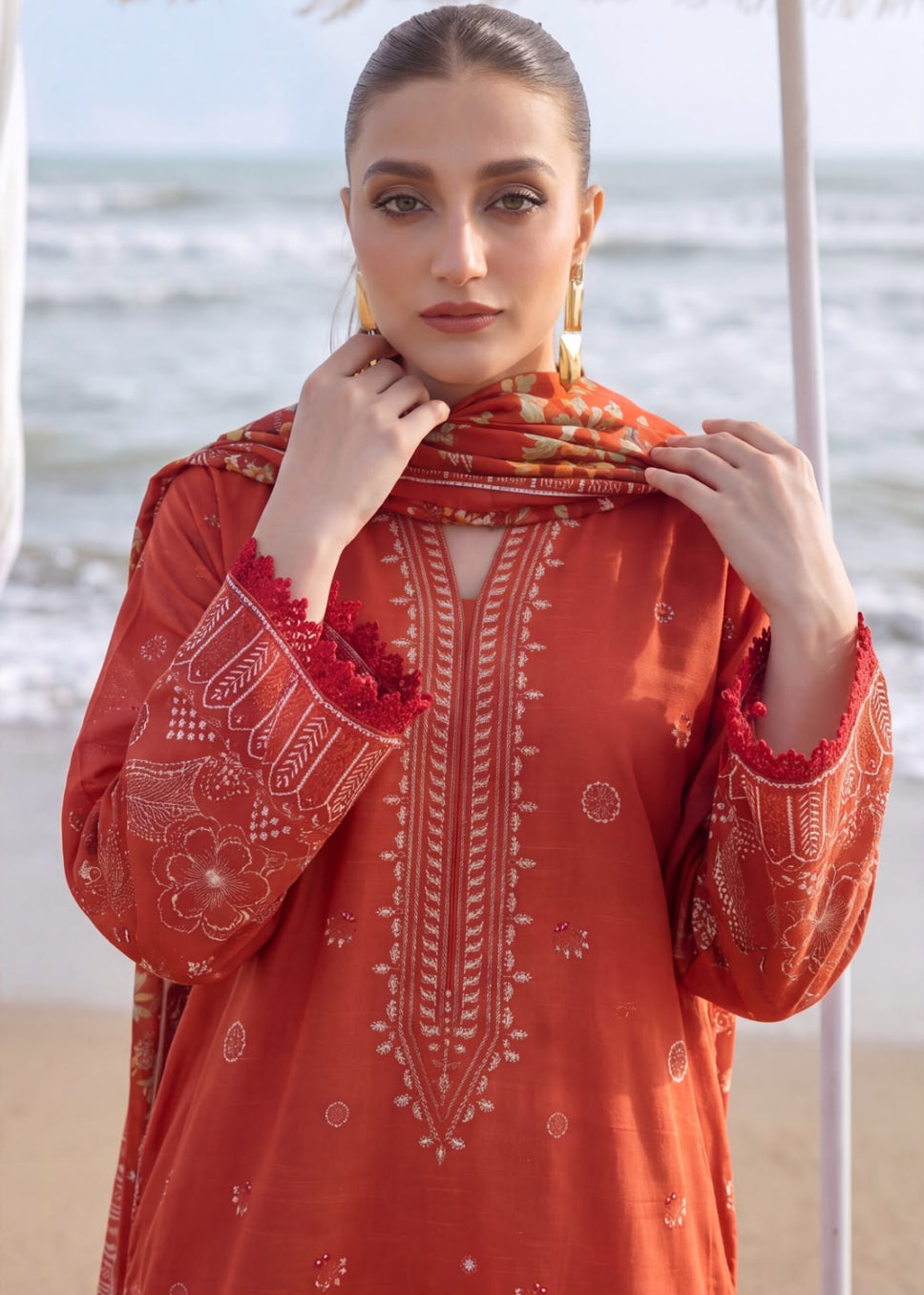 A woman wearing a rust-orange embroidered kurta with a matching dupatta, standing on a beach with the ocean in the background.