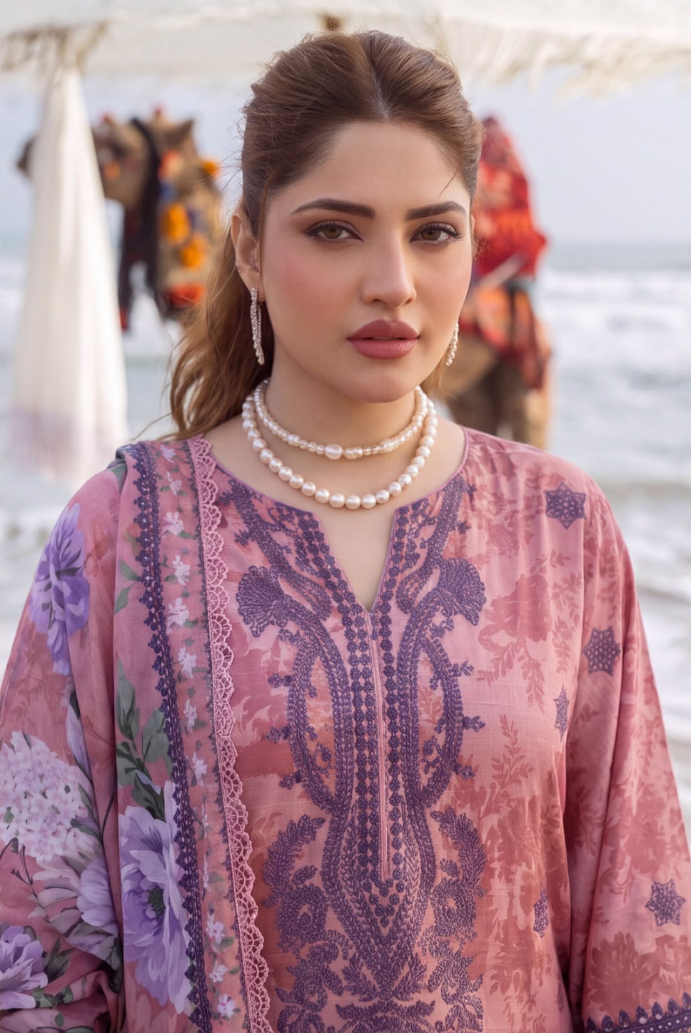 A woman wearing a pink and purple printed traditional outfit with floral patterns and lace trim, photographed outdoors with a blurred beach or water background.
