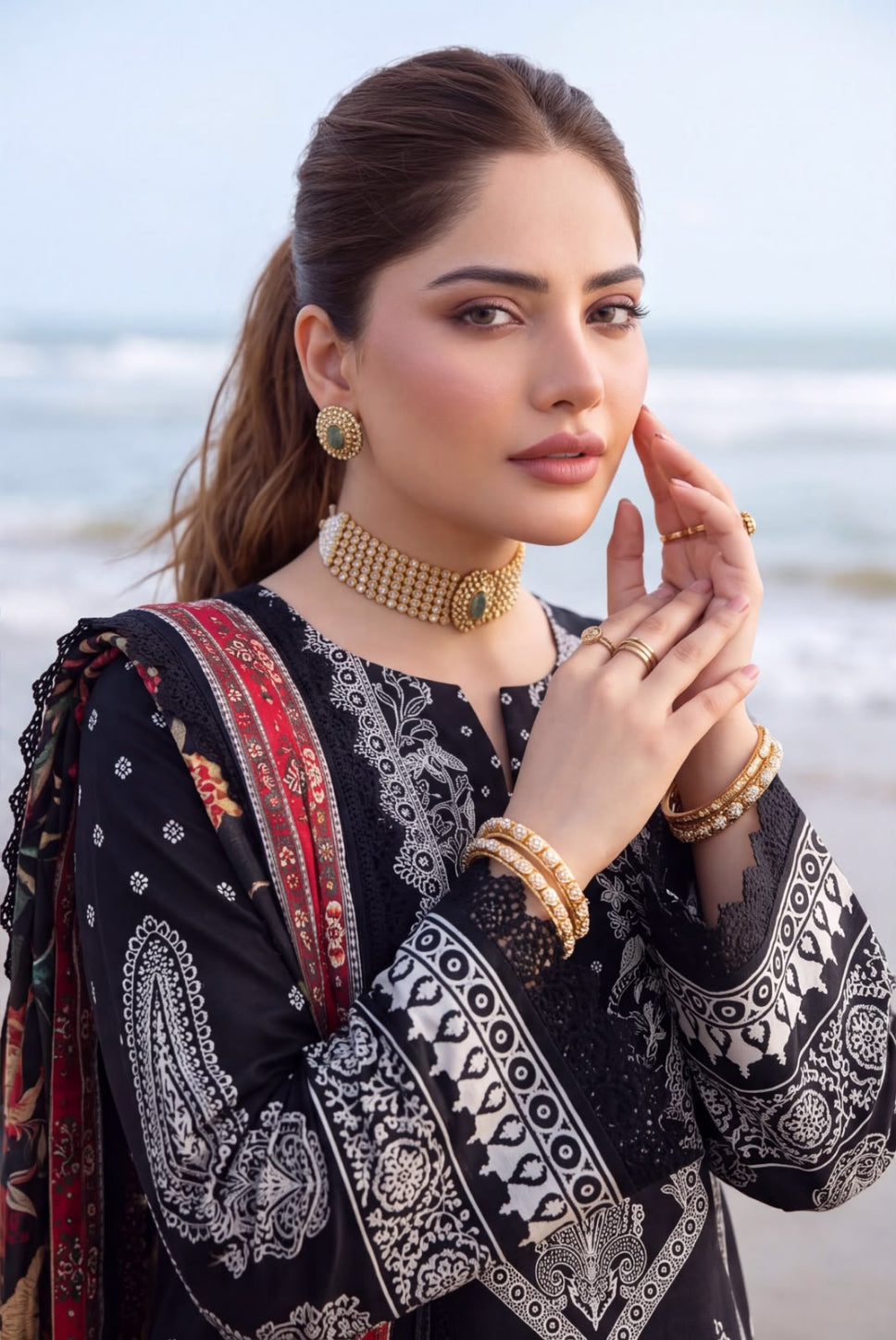 A woman posing on a beach wearing a black 3-piece salwar kameez set with white and red printed patterns, gold jewelry, and a matching black dupatta.