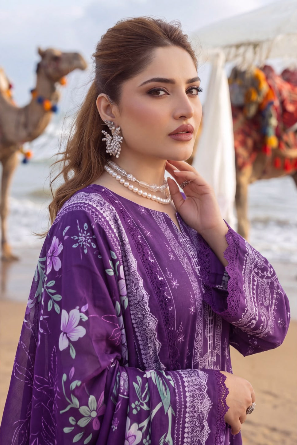 A woman wearing a purple embroidered traditional outfit with floral patterns, standing on a beach with camels and a tent blurred in the background.