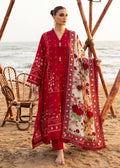 A woman stands on a sandy beach wearing a red embroidered traditional outfit with a matching printed dupatta. The outfit has intricate floral and geometric embroidery, and the dupatta features a cream base with red and pink floral motifs and a red border.