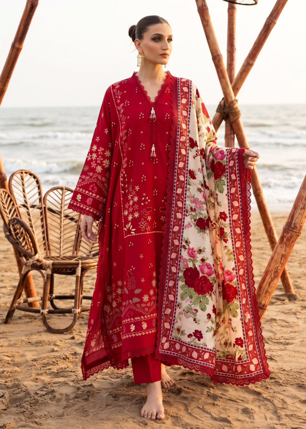 A woman stands on a sandy beach wearing a red embroidered traditional outfit with a matching printed dupatta. The outfit has intricate floral and geometric embroidery, and the dupatta features a cream base with red and pink floral motifs and a red border.