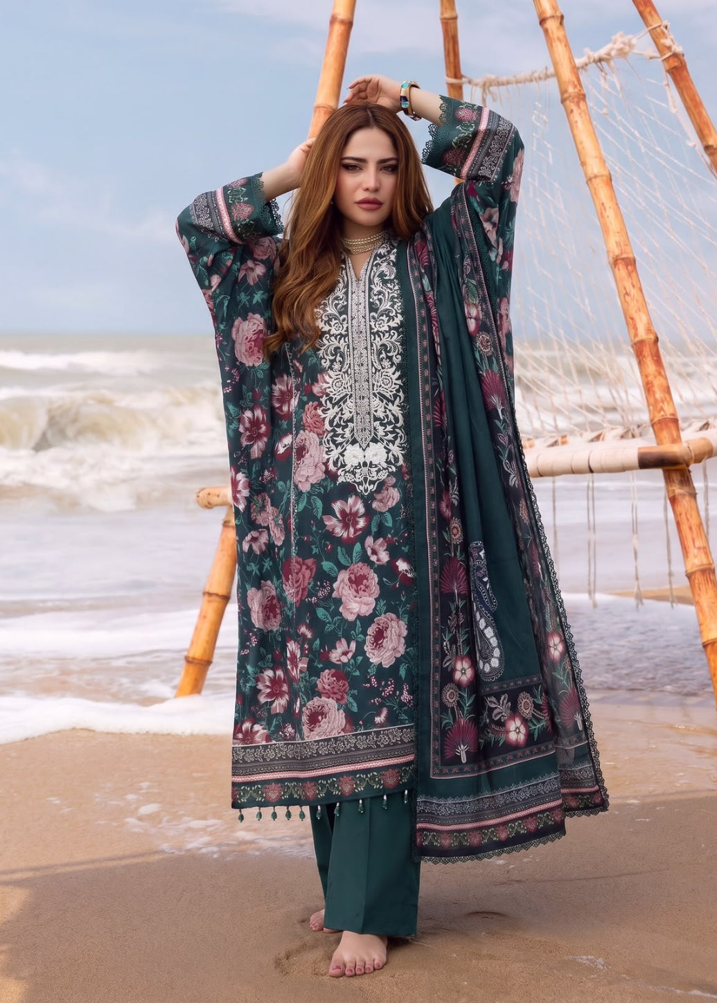 A woman stands barefoot on a sandy beach wearing a long dark teal floral embroidered kurta with matching pants and dupatta. The outfit features pink and red floral patterns with white embroidery on the chest.