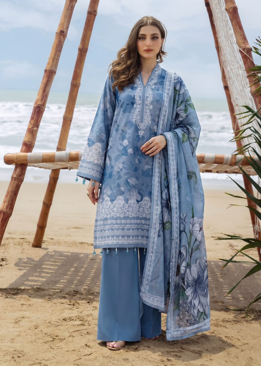 A woman stands on a sandy beach wearing a light blue printed traditional outfit with matching wide-leg pants and a draped dupatta. The outfit features white floral embroidery and blue-green floral motifs, with a bamboo beach chair and ocean in the background.