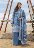 A woman stands on a sandy beach wearing a light blue printed traditional outfit with matching wide-leg pants and a draped dupatta. The outfit features white floral embroidery and blue-green floral motifs, with a bamboo beach chair and ocean in the background.
