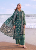 A woman stands barefoot on a sandy beach with the ocean in the background, wearing a green traditional outfit with intricate beige and white floral embroidery and a matching dupatta.