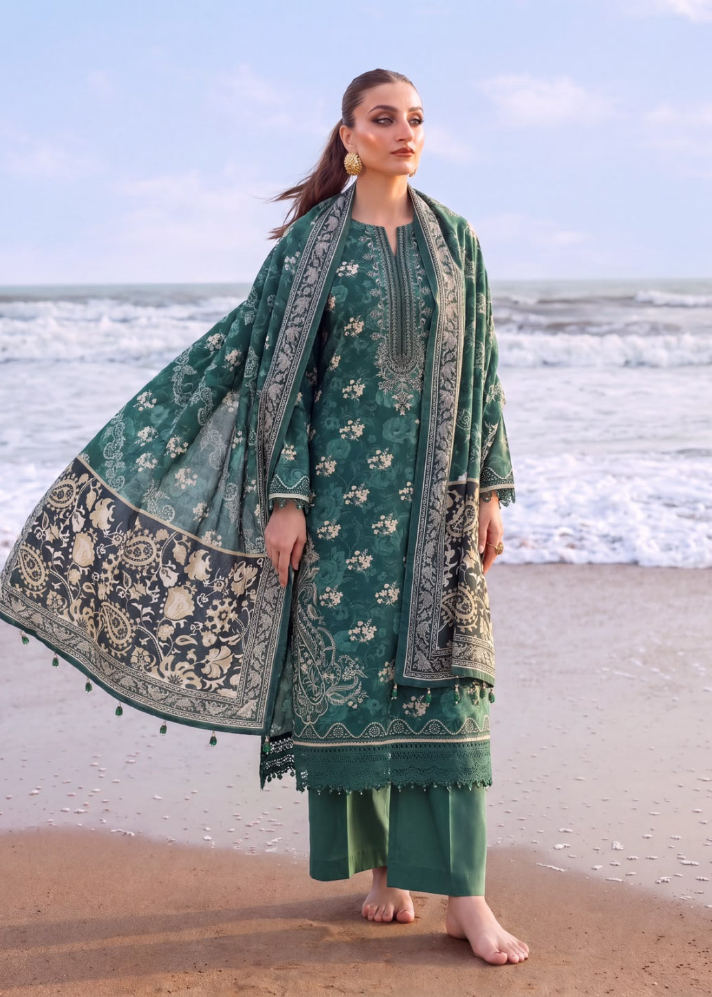 A woman stands barefoot on a sandy beach with the ocean in the background, wearing a green traditional outfit with intricate beige and white floral embroidery and a matching dupatta.