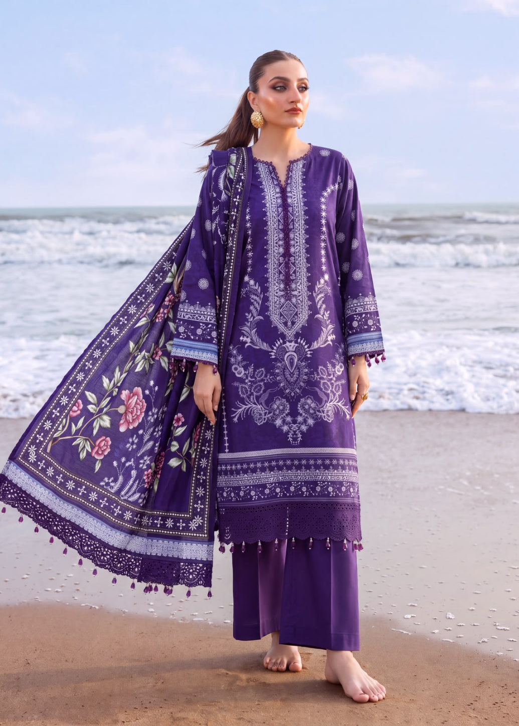 A woman stands barefoot on a sandy beach wearing a purple embroidered traditional outfit with a matching patterned dupatta. The background shows the ocean waves and a pale blue sky.