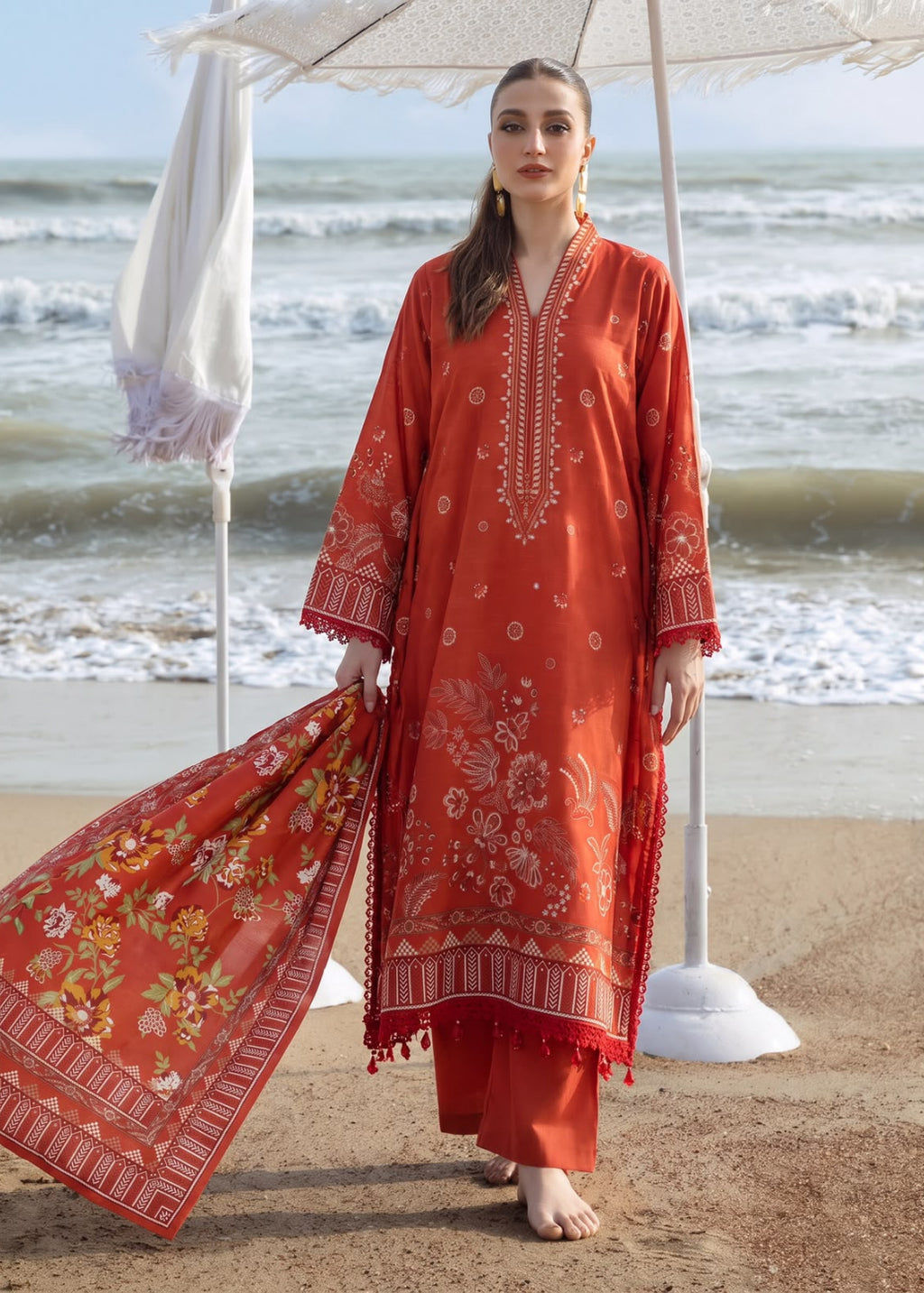 A woman stands on a sandy beach under a white parasol, wearing a rust-orange traditional embroidered kurta set with matching pants and a coordinating printed dupatta.