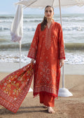 A woman stands on a sandy beach under a white parasol, wearing a rust-orange traditional embroidered kurta set with matching pants and a coordinating printed dupatta.