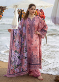 A woman stands on a beach wearing a pink traditional outfit with floral prints and dark purple embroidery, holding out a matching dupatta. Camels and umbrellas are visible in the background near the ocean.