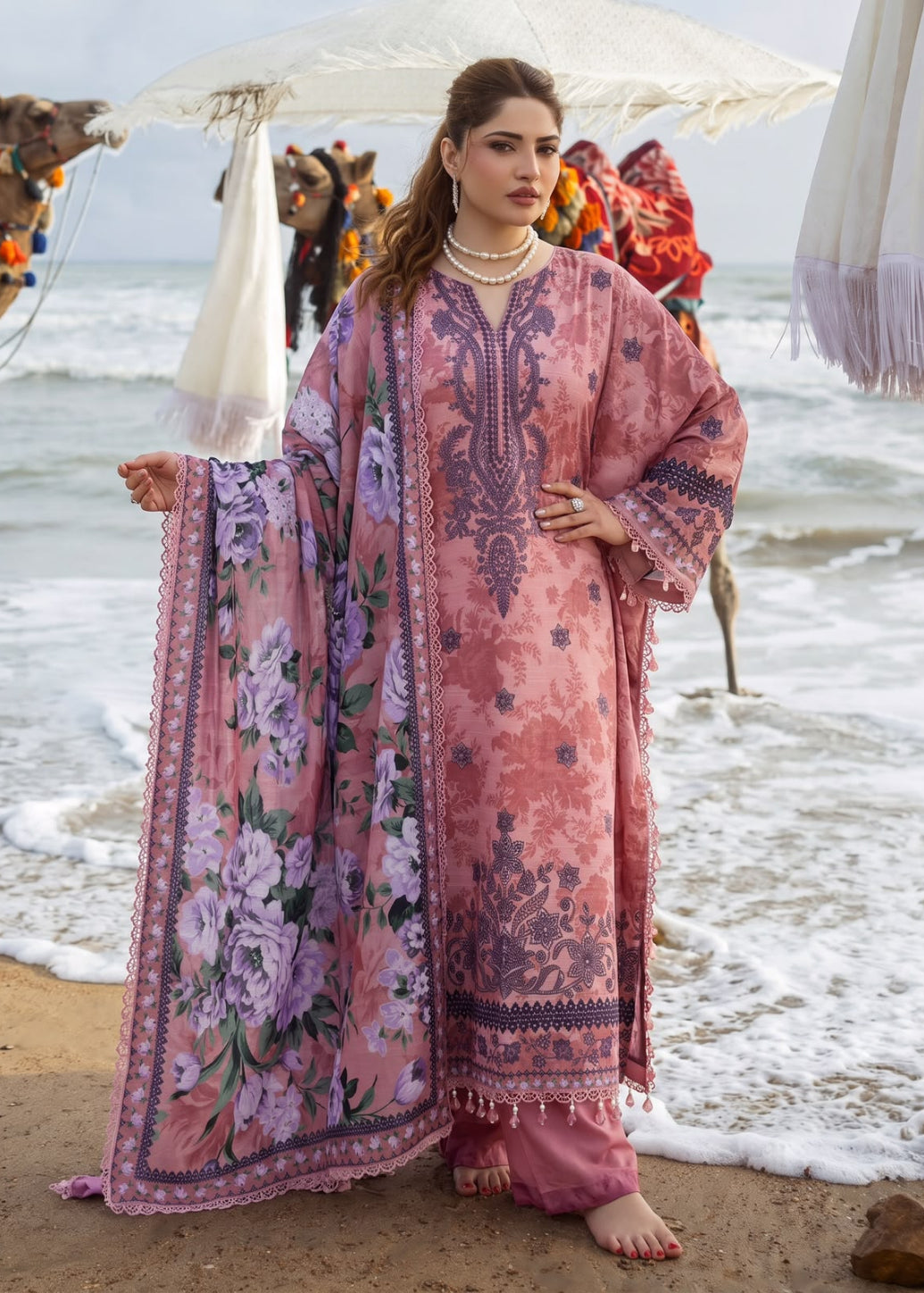 A woman stands on a beach wearing a pink traditional outfit with floral prints and dark purple embroidery, holding out a matching dupatta. Camels and umbrellas are visible in the background near the ocean.