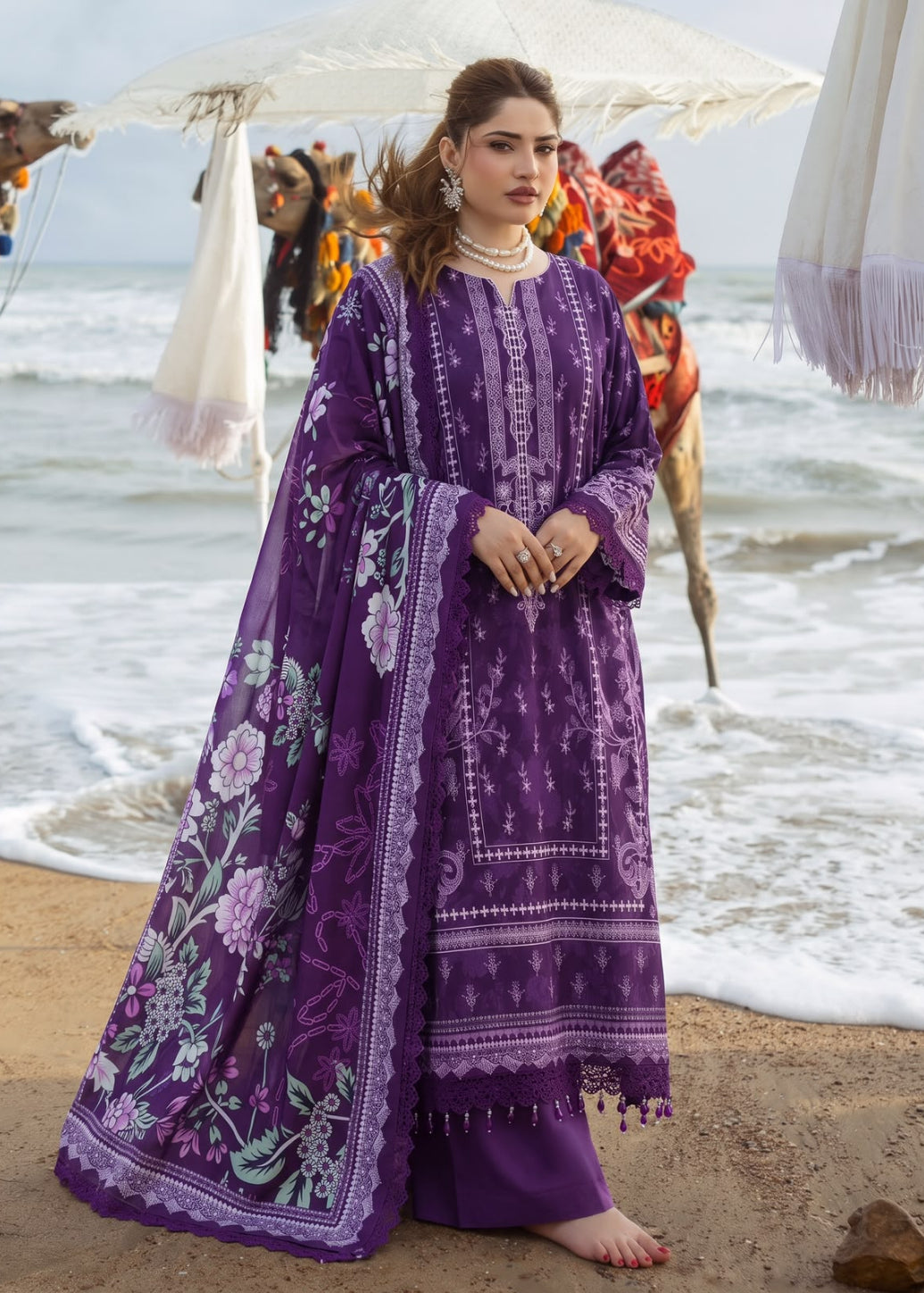 A woman stands on a sandy beach wearing a purple embroidered traditional outfit with a matching dupatta. The outfit features intricate floral and geometric patterns in shades of purple, lavender, and white.