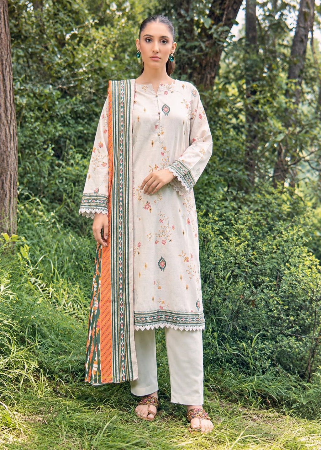 A woman stands outdoors in a lush green forest setting wearing a traditional Indian salwar kameez suit. The suit consists of a light beige kurta with delicate floral prints, matching beige trousers, and a vibrant orange dupatta with a detailed green border.