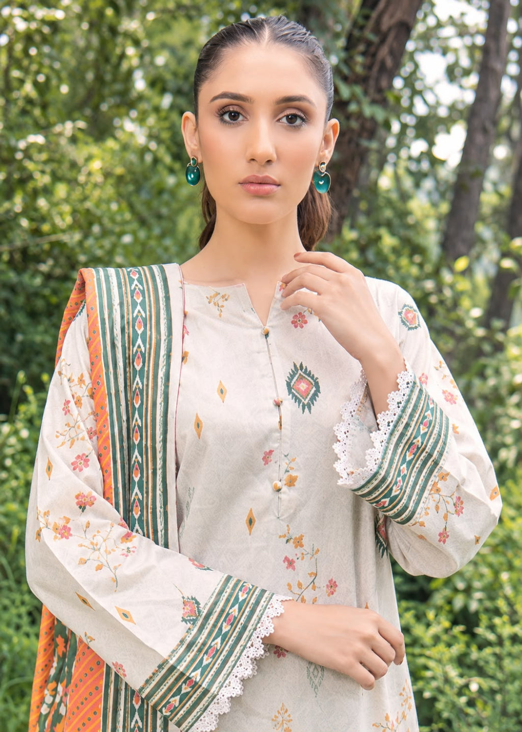 A woman wearing a traditional three-piece salwar kameez suit in off-white, featuring green and orange patterned borders, floral prints, and lace cuffs, paired with a matching dupatta, set against a blurred green forest background.
