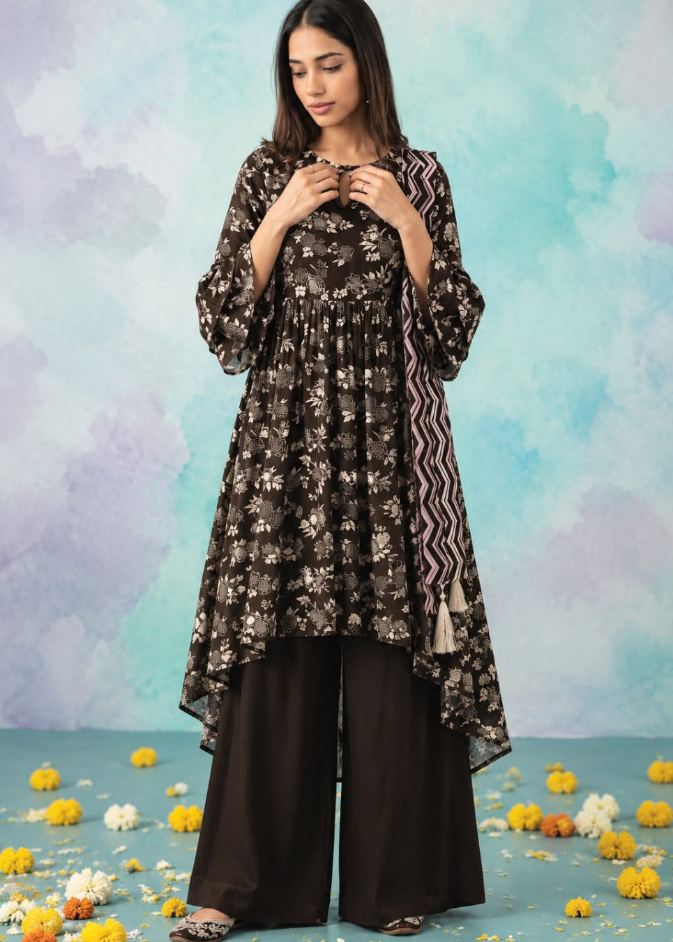 A woman models a three-piece black kurta set with a white floral print, wide-leg black trousers, and a black-and-white patterned dupatta, set against a pastel blue and purple watercolor background with scattered yellow flowers on the floor.