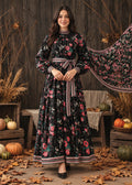 A woman stands in a rustic fall-themed setting wearing a long black floral dress with pink, red, and green floral print, a matching waist tie belt, and a matching dupatta. The background features a dark wooden wall, pumpkins, and scattered autumn leaves.