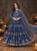 A woman models a royal blue anarkali-style dress with beige floral prints and a matching patterned dupatta, posed in front of a rustic wooden wall decorated with pumpkins and autumn leaves.