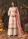 A woman stands outdoors against a rustic wooden wall, wearing a cream-colored traditional outfit with pink floral embroidery and a matching pink dupatta. The scene includes pumpkins, dry grass, and scattered autumn leaves.