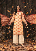 A woman stands outdoors in front of a dark wooden wall with scattered autumn leaves, wearing a peach kurta with orange polka dots, wide cream pants, and a rust-colored patterned dupatta spread wide on both sides.