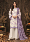 A woman stands in a rustic outdoor-style setting with a dark wood plank wall, autumn leaves on the ground, and decorative pumpkins and dried plants. She is wearing a traditional three-piece outfit: a cream-colored kurta with purple floral embroidery and a purple border, white wide-leg pants, and a matching purple dupatta.