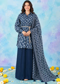 A woman modeling a navy blue three-piece ethnic suit set, featuring a long-sleeve button-up kurta with a white floral print, matching navy wide-leg pants, and a navy printed dupatta with a geometric pattern and tassel border, set against a blue and purple watercolor background with yellow flowers on the floor.