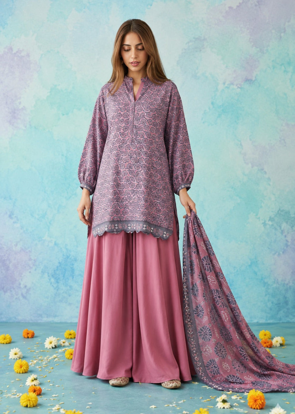 A woman models a traditional three-piece salwar kameez set in dusty purple and pink. The kurta features a purple floral pattern, paired with wide-leg pink trousers and a matching patterned dupatta, set against a textured blue and purple backdrop with scattered yellow flowers.