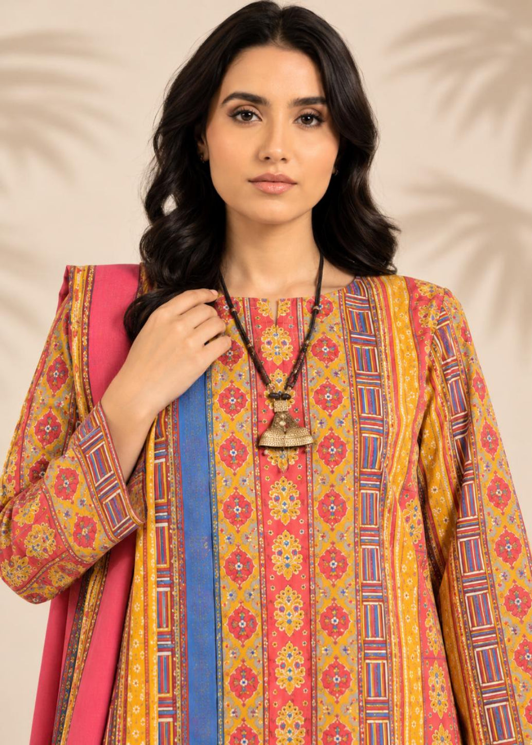 A woman modeling a vibrant three-piece Indian ethnic suit set featuring a multicolor printed kurta in yellow, orange, and red with blue accents, a solid pink dupatta, and a brown beaded necklace.