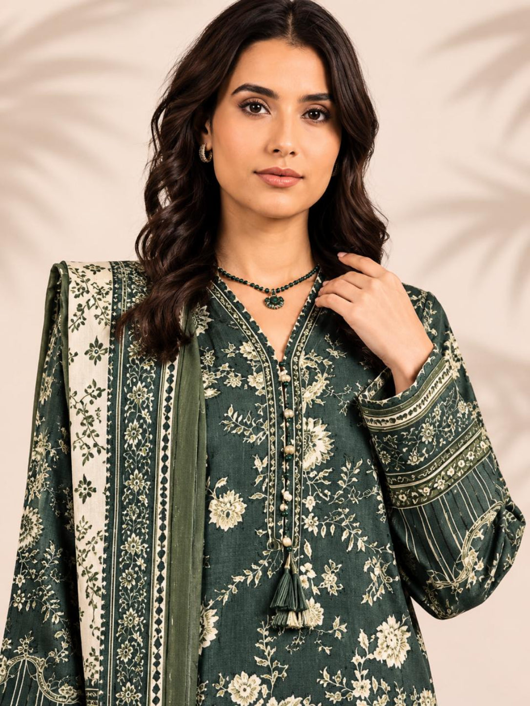 A woman modeling a dark green floral printed kurta with a matching dupatta, featuring cream-colored floral motifs and detailed embroidery on the sleeves, set against a soft beige background with leafy shadows.