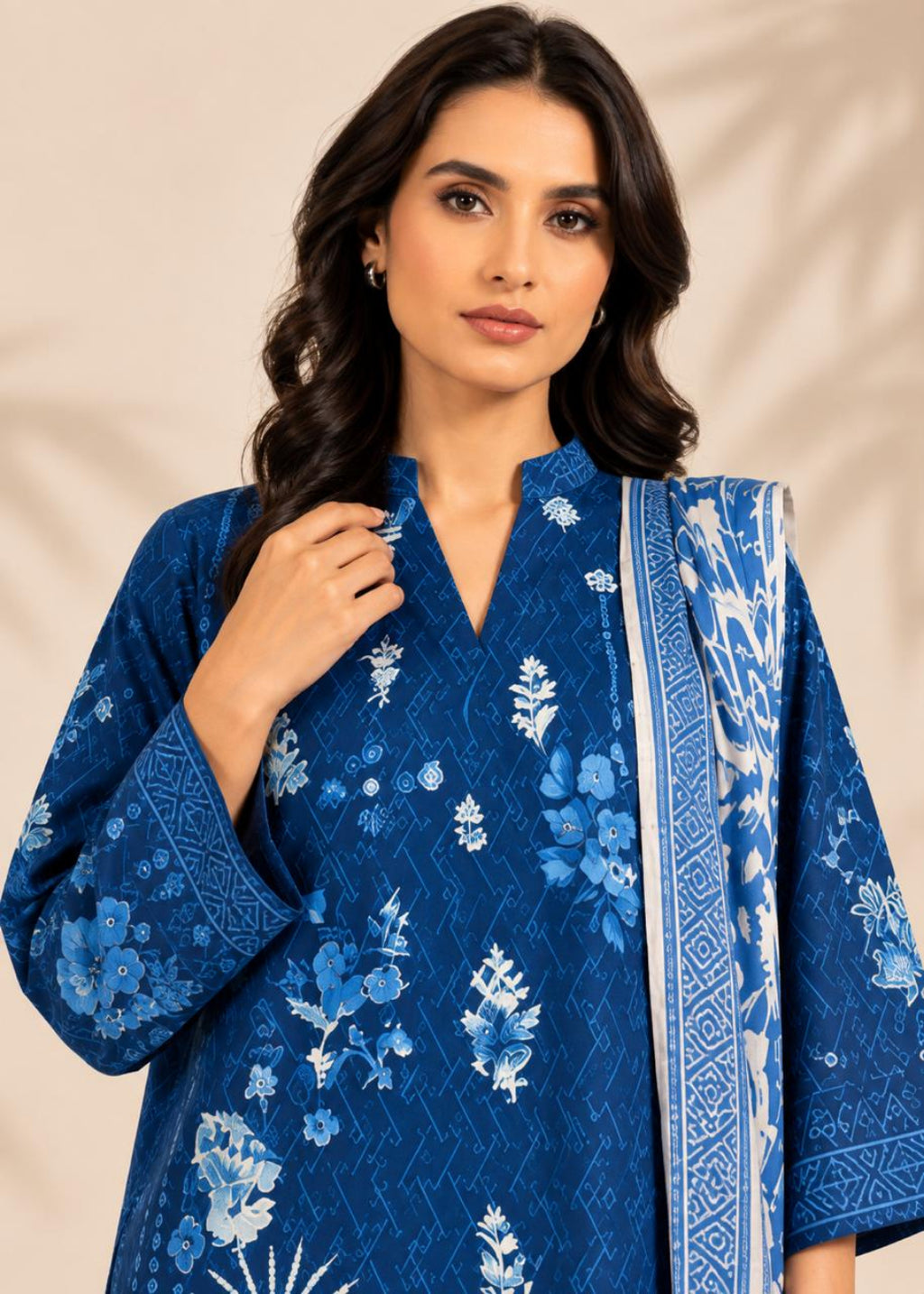 A woman wearing a royal blue embroidered kurta with white floral patterns and a matching patterned dupatta, set against a warm beige background with soft leaf shadows.
