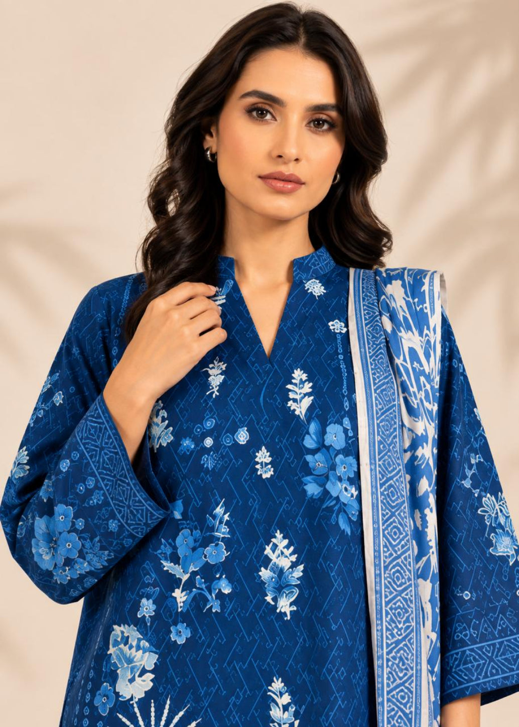 A woman wearing a royal blue embroidered kurta with white floral patterns and a matching patterned dupatta, set against a warm beige background with soft leaf shadows.