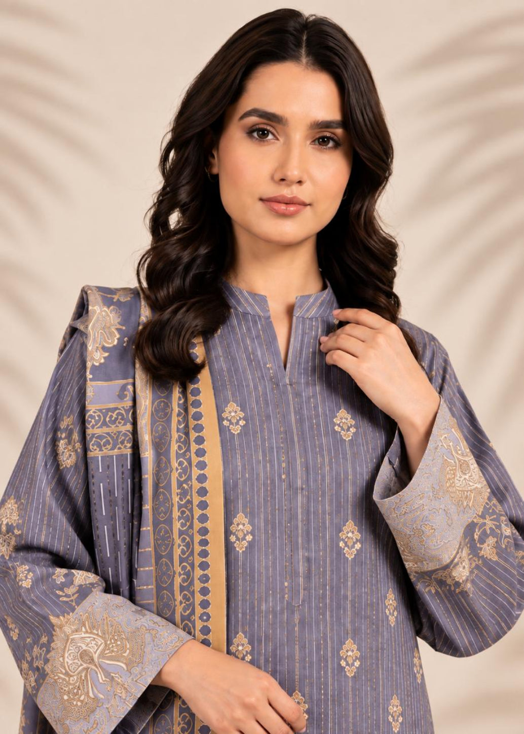 A woman modeling a lavender-blue and gold embroidered traditional kurta set against a warm beige background with subtle leaf patterns.