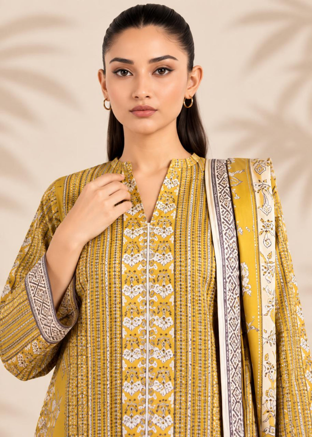 A woman modeling a mustard yellow and white printed three-piece salwar kameez suit with a matching dupatta, set against a warm beige background with subtle leaf shadows.