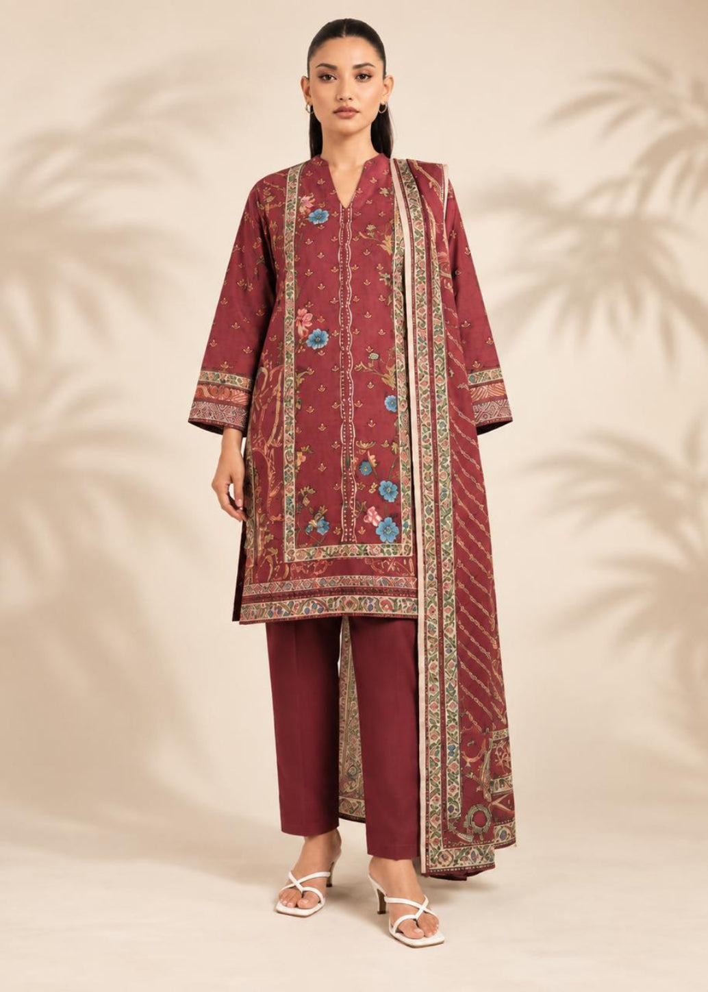 A woman models a three-piece traditional outfit consisting of a maroon kurta with colorful floral embroidery, matching maroon trousers, and a coordinating embroidered dupatta, set against a warm beige background with soft palm leaf shadows.