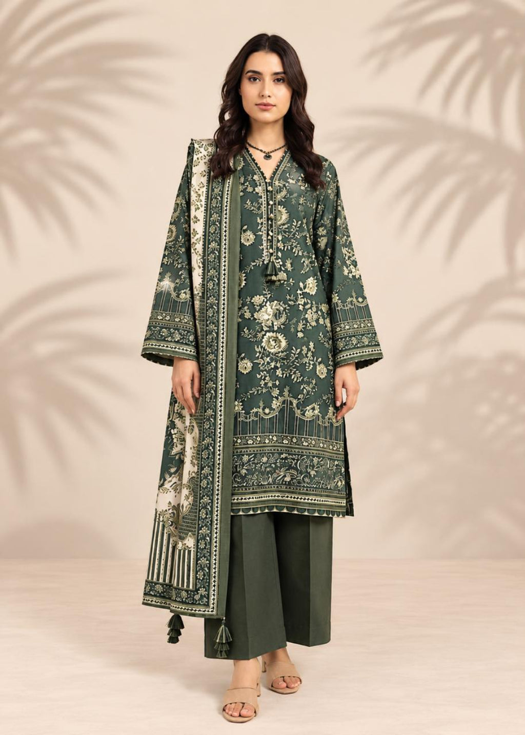 A woman modeling a dark green three-piece traditional outfit with beige floral prints, featuring a long kurta, wide sleeves, matching trousers, and a coordinating dupatta, set against a warm beige background with palm leaf shadows.
