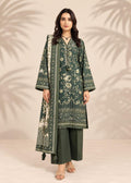 A woman modeling a dark green three-piece traditional outfit with beige floral prints, featuring a long kurta, wide sleeves, matching trousers, and a coordinating dupatta, set against a warm beige background with palm leaf shadows.