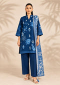 A woman models a three-piece blue ethnic suit set featuring a floral printed kurta, matching trousers, and a patterned dupatta, set against a beige background with soft palm leaf shadows.