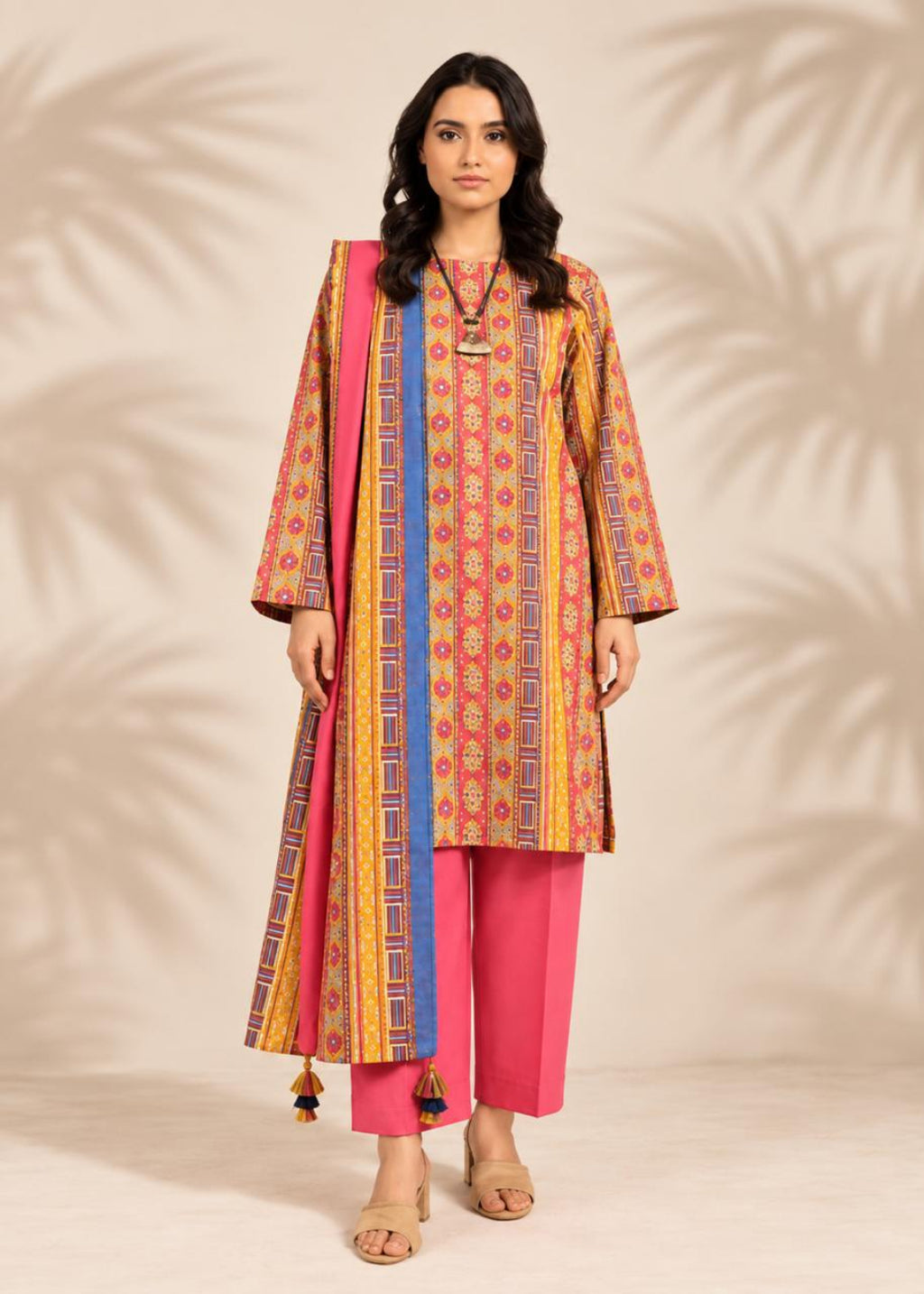 A woman modeling a traditional Indian outfit consisting of a long printed kurta in orange, yellow, and blue, paired with bright pink pants and a pink dupatta with blue borders, set against a beige background with leaf shadows.