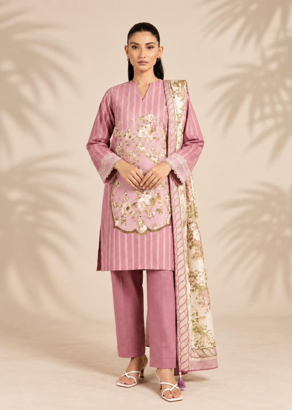 A woman models a traditional three-piece salwar kameez suit in dusty pink, featuring vertical stripes, floral embroidery on the chest, matching trousers, and a patterned dupatta, set against a beige background with soft palm leaf shadows.