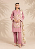 A woman models a traditional three-piece salwar kameez suit in dusty pink, featuring vertical stripes, floral embroidery on the chest, matching trousers, and a patterned dupatta, set against a beige background with soft palm leaf shadows.