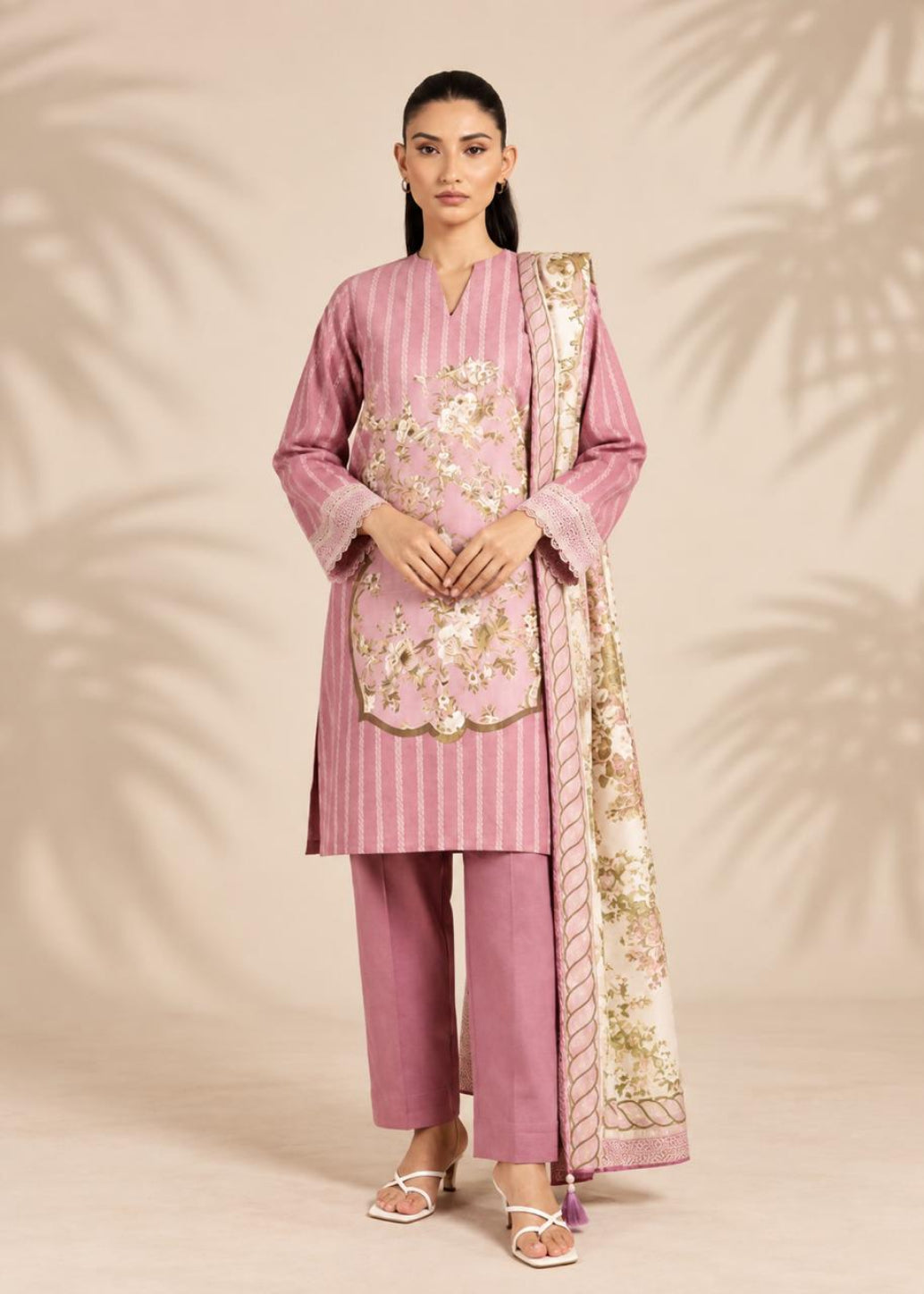 A woman models a traditional three-piece salwar kameez suit in dusty pink, featuring vertical stripes, floral embroidery on the chest, matching trousers, and a patterned dupatta, set against a beige background with soft palm leaf shadows.